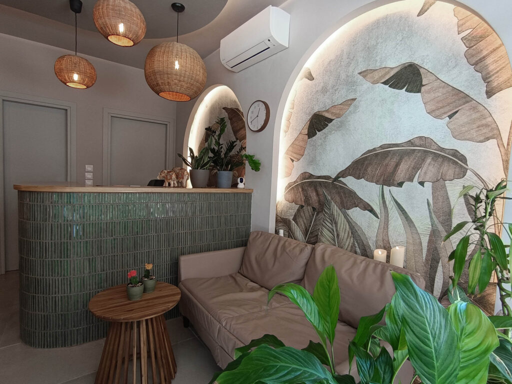 Φωτογραφία του χώρου του καθιστικού του Nature Thai Massage. Μπροστά από τον πάγκο της reception υπάρχει καναπές και στον τοίχο σχηματίζονται δύο καμάρες με την χαρακτηριστική ταπετσαρία με τα τροπικά φύλλα και τον κρυφό φωτισμό.